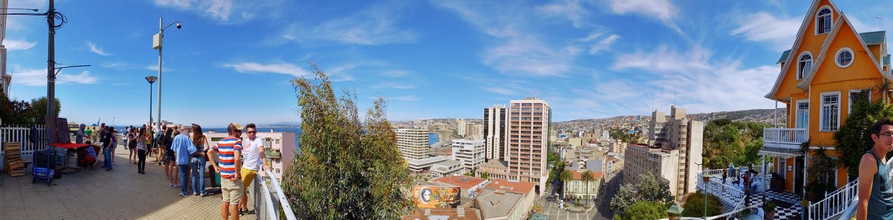 Panoramic view of Valparaíso, Chile from an elevated viewpoint showing tourists on a paved walkway with white railing, colorful Victorian houses on hillsides including a prominent orange building, dense urban cityscape below, and Pacific Ocean visible on the horizon under blue sky with wispy clouds