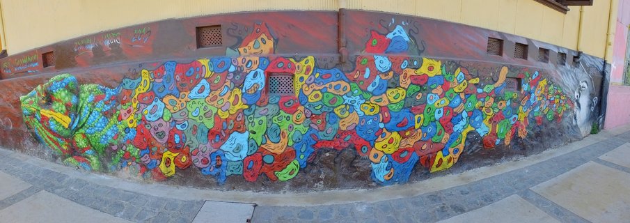 Colorful street art mural on a curved wall featuring a green chameleon on the left, abstract multicolored organic shapes in the center, and a grayscale portrait on the right, with a yellow building above and paved sidewalk below