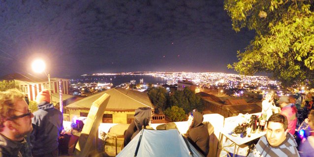 Night photograph showing people gathered at a viewpoint with camping tent, overlooking an illuminated coastal city and bay during New Year's Eve celebration in Chile