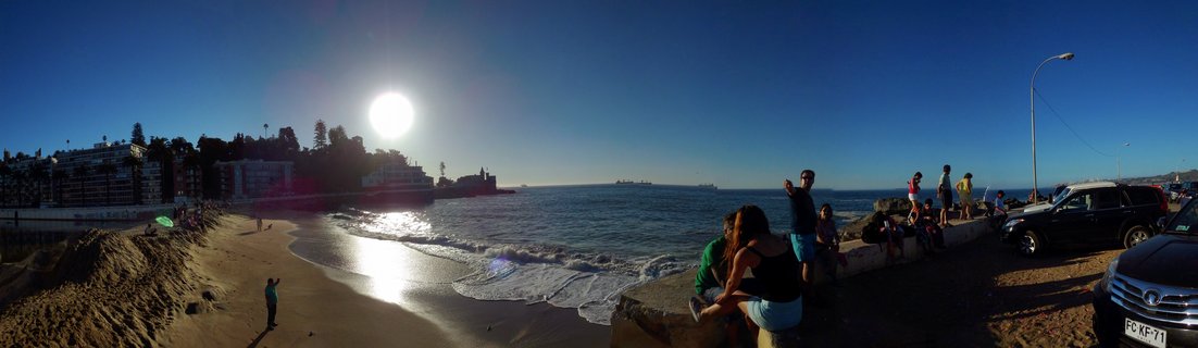 Wide coastal panorama showing people sitting on a concrete seawall beside a sandy beach, with the bright sun over the ocean, buildings on hillsides, and parked cars nearby