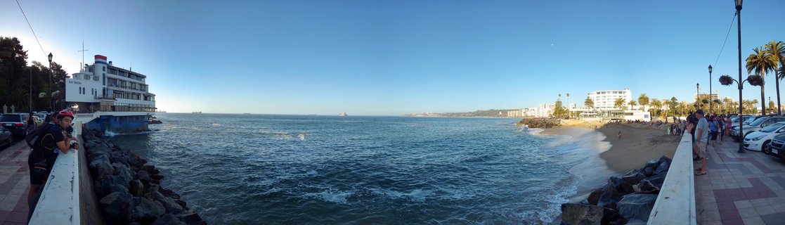 Panoramic coastal scene showing a white yacht club building on a pier, Pacific Ocean with waves, sandy beach, promenade with people walking, palm trees, and clear blue sky in Viña del Mar, Chile