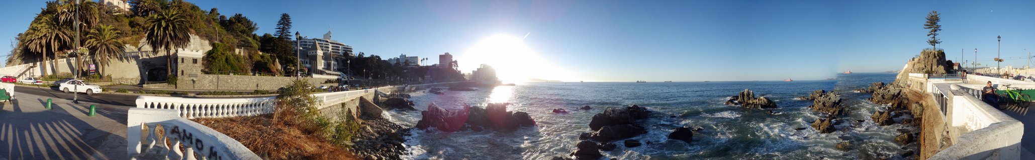 Wide panoramic photograph of Chilean Pacific coast at sunset showing rocky shoreline, ocean waves, white decorative railings, palm trees, hillside buildings, and bright sun over water