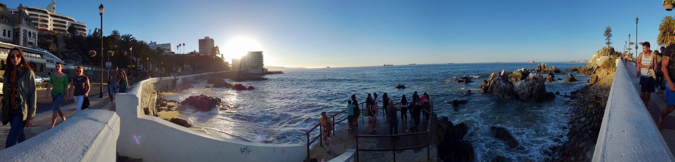 People walking on a coastal promenade with white walls overlooking the Pacific Ocean at sunset, with rocky formations in the water and buildings on the hillside