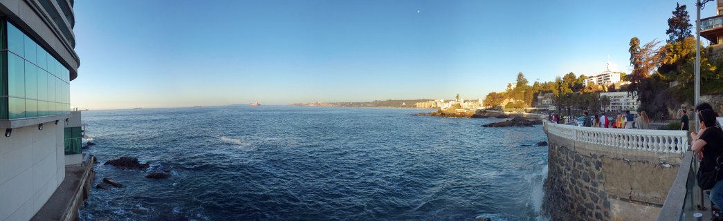 Panoramic coastal view showing Pacific Ocean, stone seawall promenade with people walking, modern glass building on left, hillside with trees and white buildings on right, clear blue sky