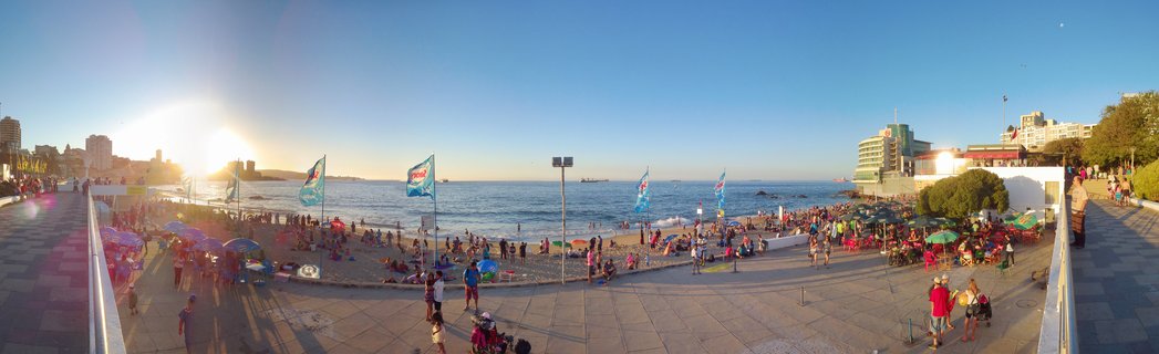 Wide panoramic beach scene at sunset showing crowded shoreline with blue flags, boardwalk with people walking, ocean waves, and coastal buildings under clear blue sky with golden sunlight