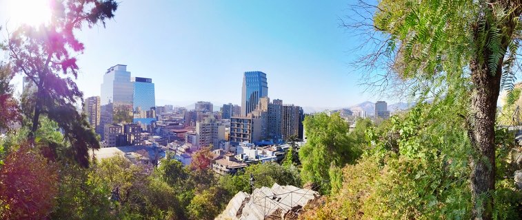 City skyline of Santiago Chile with modern glass skyscrapers, green trees in foreground, and mountains visible in the distance under blue sky with sun flare