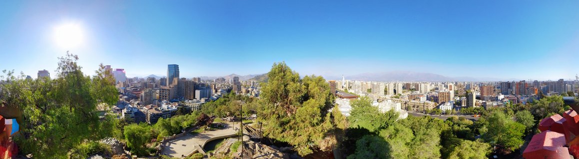 Wide cityscape view of Santiago de Chile showing urban buildings, green trees in foreground, and Andes mountains in the distance under bright blue sky with visible sun