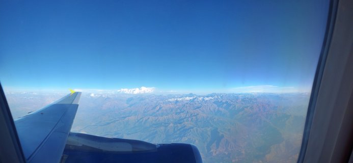 Aerial view from airplane window showing aircraft wing with yellow wingtip and mountain range with snow-capped peaks under clear blue sky