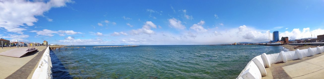 Wide waterfront panorama of Puerto Natales, Chile showing concrete breakwaters framing turquoise harbor water with cityscape and blue cylindrical building under cloudy sky