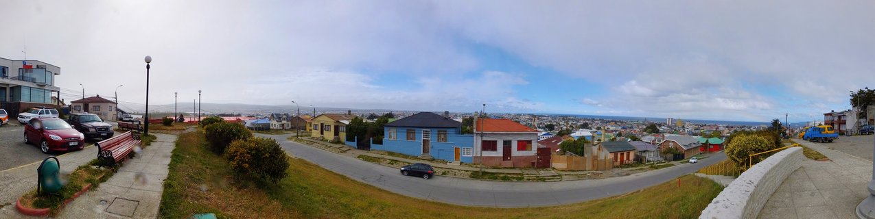 Wide panoramic photograph showing a curved residential street in Puerto Natales, Chile, with colorful blue, red, and yellow houses, parked cars, and distant water view under cloudy sky