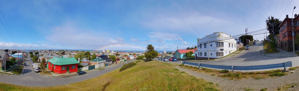 Panoramic street view of colorful houses on grassy hills in Puerto Natales, Chile, with blue sky and distant water visible