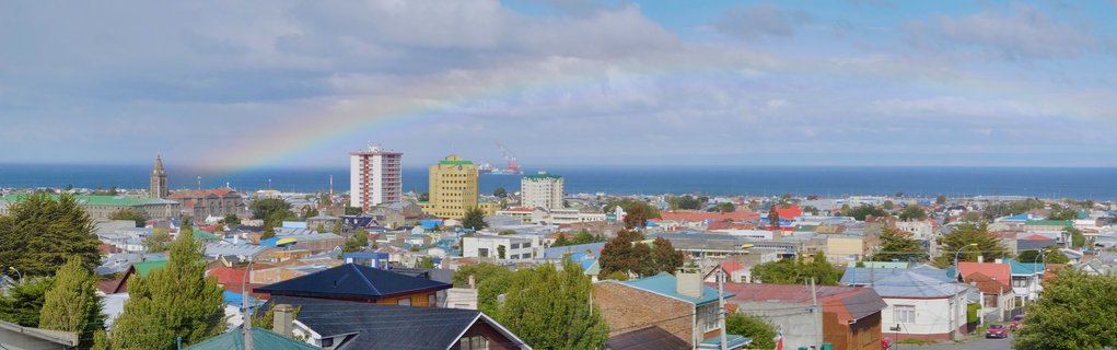 Panoramic view of Puerto Natales, Chile showing a rainbow over the city with colorful buildings, church tower, trees, and the ocean in the background under cloudy sky
