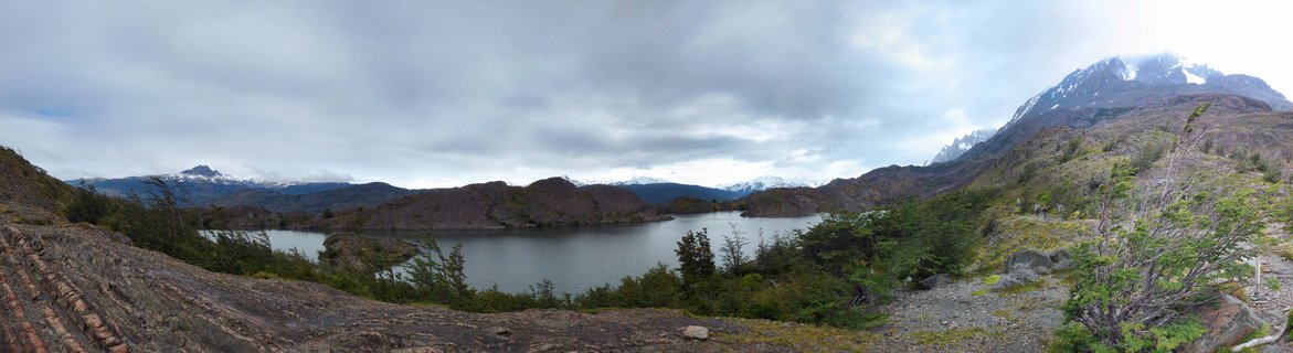 Panoramic landscape of Torres del Paine National Park showing a lake surrounded by rocky terrain, native vegetation, and snow-capped mountains under cloudy skies