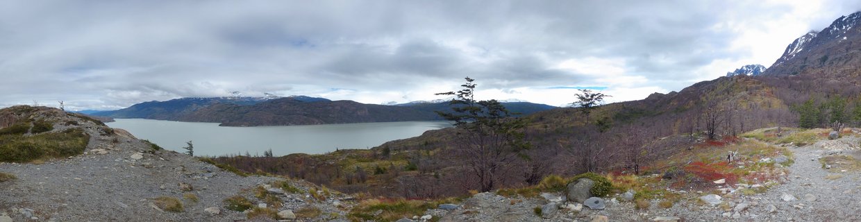 Wide landscape photograph showing a lake surrounded by mountains in Torres del Paine National Park, Chile, with rocky terrain and sparse trees in the foreground under cloudy skies
