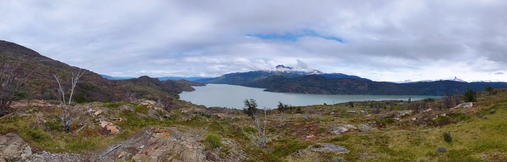 Wide landscape showing a glacial lake with gray-green water surrounded by rocky hills with sparse vegetation, snow-capped mountains visible in the distance under cloudy sky