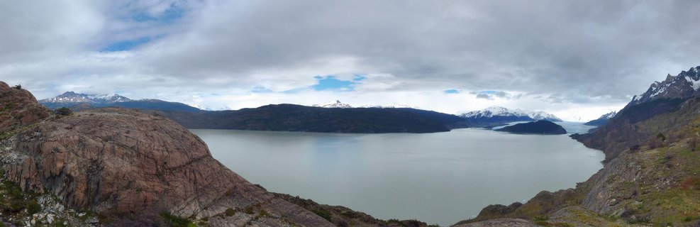 Wide landscape view of Torres del Paine National Park showing a large glacial lake surrounded by rocky mountains with snow-capped peaks under cloudy sky