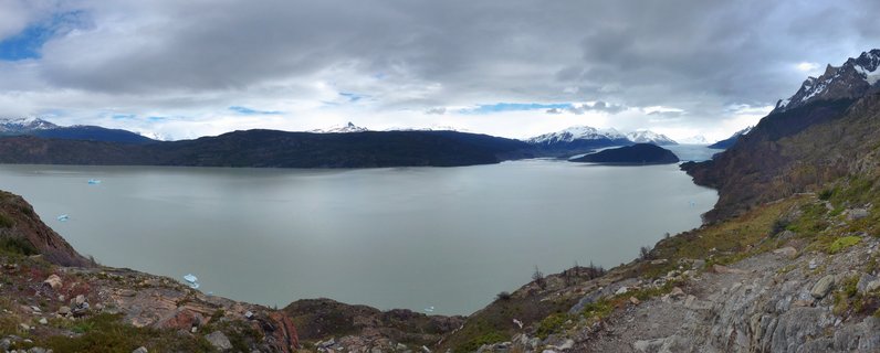 Wide landscape view of a glacial lake with floating icebergs, surrounded by rocky terrain in the foreground and snow-capped mountains in the distance under cloudy skies in Torres del Paine, Chile