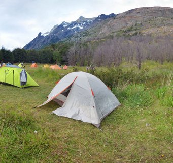 Multiple colorful camping tents in a green meadow with snow-capped mountains and rocky peaks in the background at Torres del Paine National Park