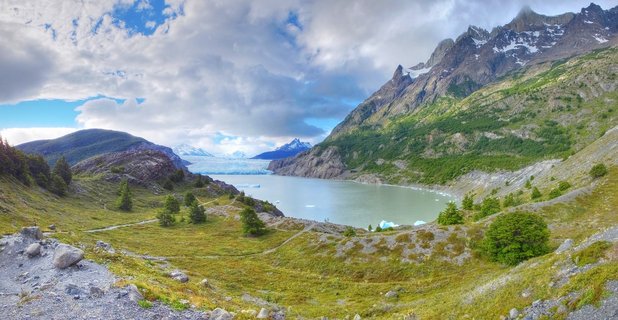 Panoramic landscape showing Grey Lake with floating icebergs, surrounded by green hillsides and dramatic snow-capped granite mountain peaks of Torres del Paine National Park in Chile