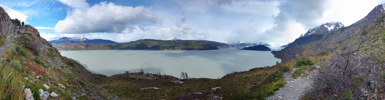Panoramic landscape showing a large grey glacial lake surrounded by mountains with snow-capped peaks, hiking trails, and cloudy sky in Torres del Paine National Park, Chile