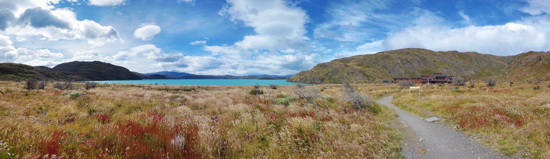 Wide landscape showing a bright turquoise lake framed by brown-green hills under a blue sky with white clouds, with a gravel path leading through grassy foreground toward buildings