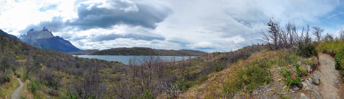 Panoramic landscape showing Torres del Paine mountain peaks on the left, a glacial lake in the center, and hiking trails through scrub vegetation in the foreground under cloudy skies