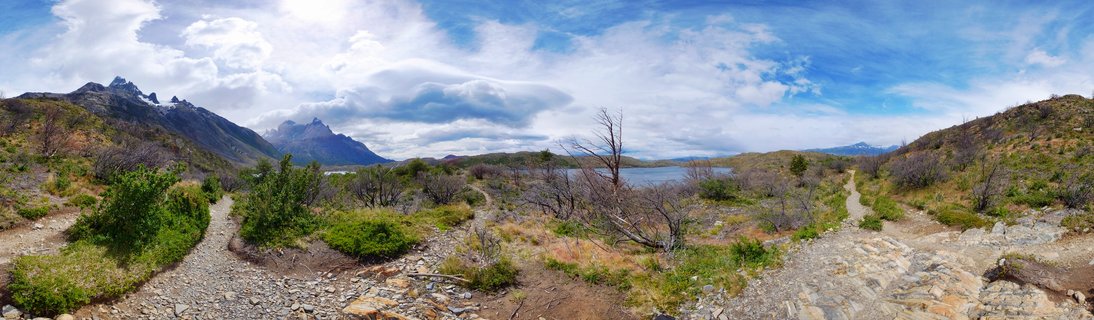 Panoramic landscape of Torres del Paine National Park showing mountain peaks, a lake, hiking trails, and shrub-covered terrain under cloudy skies