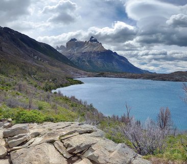 Granite mountain peaks of Torres del Paine rising above a blue glacial lake with rocky foreground and cloudy sky in Chilean Patagonia