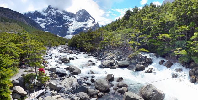 A wide mountain river with white water rapids flowing over boulders through green forest, with snow-capped peaks in the background and hikers visible on a trail at left