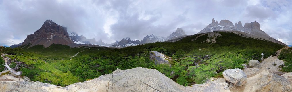 Panoramic view of Torres del Paine mountain range in Chile showing granite tower peaks, snow-capped mountains, green forest, and rocky foreground under cloudy sky