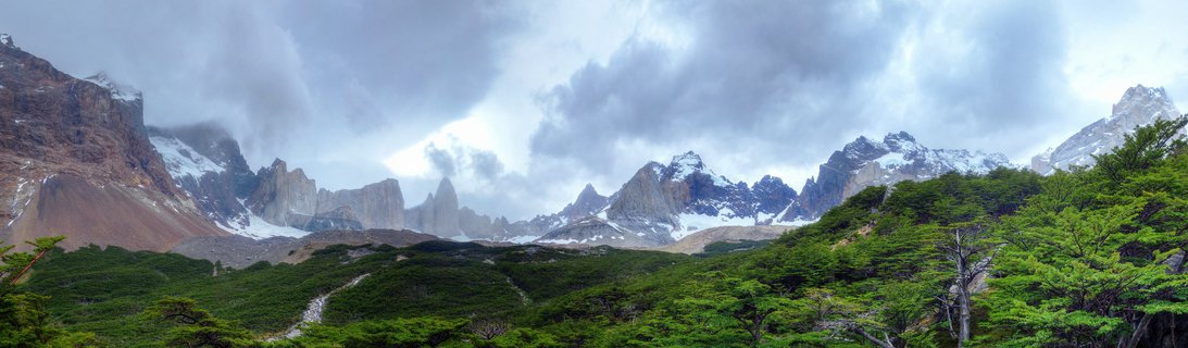 Panoramic view of Torres del Paine mountain range with snow-capped granite peaks, green forest foreground, and cloudy sky in Chilean Patagonia