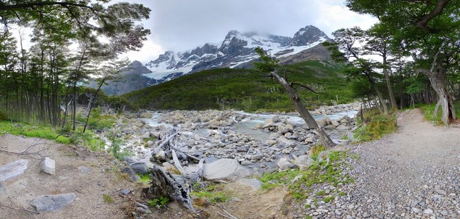 Wide landscape showing a rocky glacial river flowing through green vegetation with snow-capped mountain peaks in the background under cloudy sky, Torres del Paine, Chile
