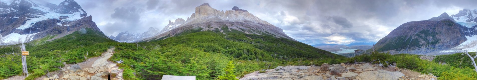 Panoramic view of Torres del Paine's three granite towers rising above green forest with hiking trail in foreground and dramatic cloudy sky