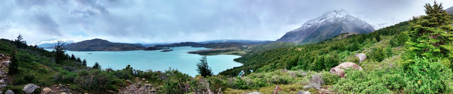 Panoramic landscape of Torres del Paine National Park showing turquoise glacial lake surrounded by green forest and snow-capped granite mountain peaks under cloudy sky