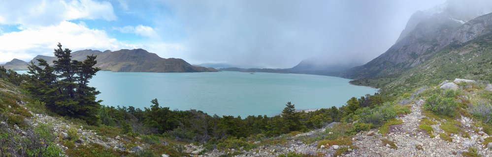 Panoramic view of turquoise Lago Pehoé glacial lake surrounded by mist-covered granite mountains of Paine Massif in Torres del Paine National Park, Chile. Dark green evergreen trees frame the left side while rocky Patagonian terrain occupies the foreground.