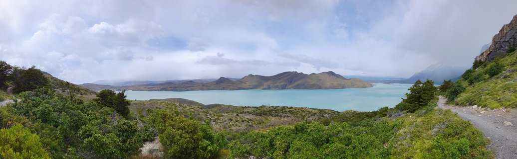 Panoramic landscape showing a turquoise glacial lake surrounded by green Patagonian vegetation in the foreground and brown mountain peaks in the distance under cloudy skies, with a hiking trail visible on the right side