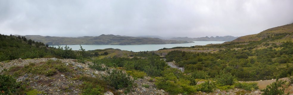 Panoramic landscape showing a turquoise glacial lake surrounded by green and brown rolling hills under an overcast sky in Torres del Paine, Chile