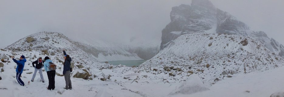 Four people in winter clothing standing on snowy rocky ground photographing a foggy mountain peak with a turquoise lake visible in the distance at Torres del Paine National Park in Chile