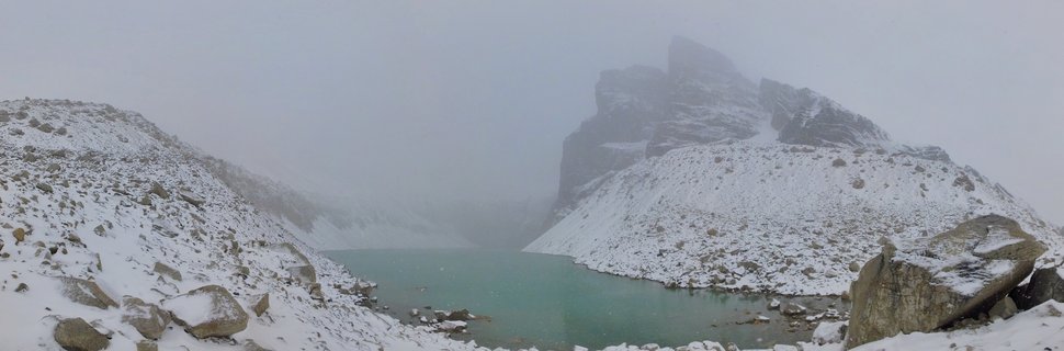 Snow-covered rocky mountains with a prominent granite tower formation rising above a turquoise alpine lake under foggy overcast sky