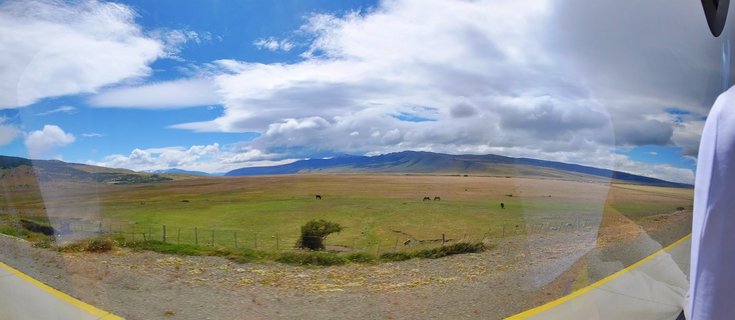 Wide landscape showing horses grazing on green and brown grasslands with mountains in the distance under a blue sky with white clouds