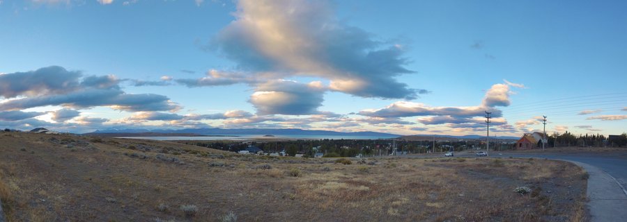 Wide panoramic landscape of Patagonian steppe with dry grassland foreground, a lake and distant mountains under a blue sky with scattered clouds, near Perito Moreno, Argentina