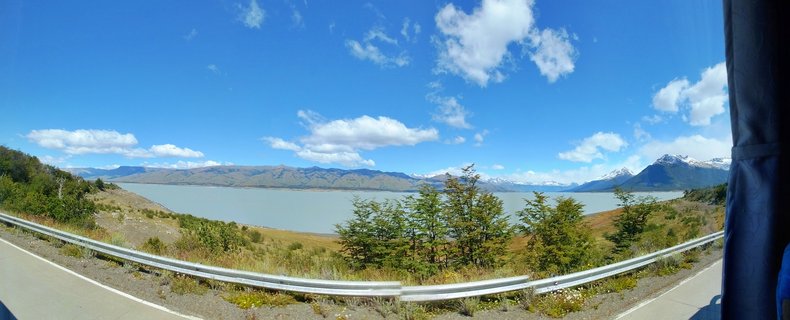 Wide landscape photograph showing a glacial lake with mountains in the background, green trees and shrubs in the middle ground, and a road with metal guardrail in the foreground under a blue sky with white clouds