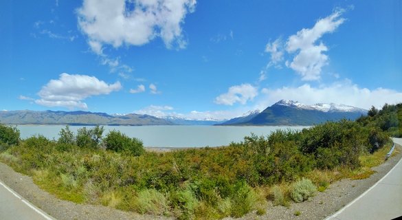 Wide landscape showing a glacial lake with snow-capped mountains in the distance, green shrubs in the foreground, and blue sky with white clouds above Perito Moreno region in Argentina