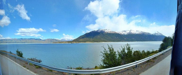 Wide landscape view of Lago Argentino with turquoise glacial water, snow-capped Andes mountains in the background, green vegetation and guardrail along roadside in foreground, blue sky with white clouds above