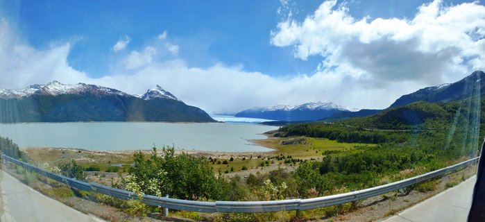 Wide landscape showing Perito Moreno Glacier in the distance across Lago Argentino, with snow-capped Andes mountains, green Patagonian vegetation in foreground, and blue sky with white clouds