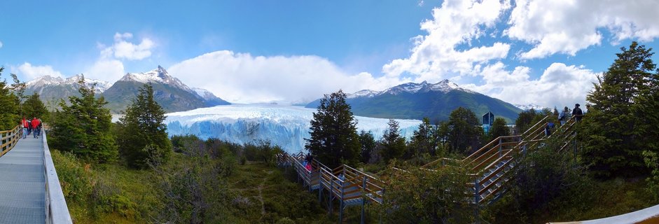 Panoramic view of Perito Moreno Glacier with wooden boardwalks and tourists in foreground, snow-capped mountains and blue sky with clouds in background, Patagonia Argentina