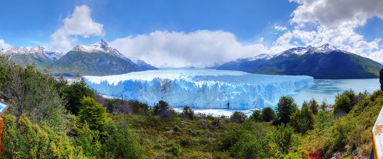 Panoramic view of Perito Moreno Glacier showing massive blue ice walls extending across a lake with snow-capped mountains in the background and green vegetation in the foreground under a partly cloudy blue sky