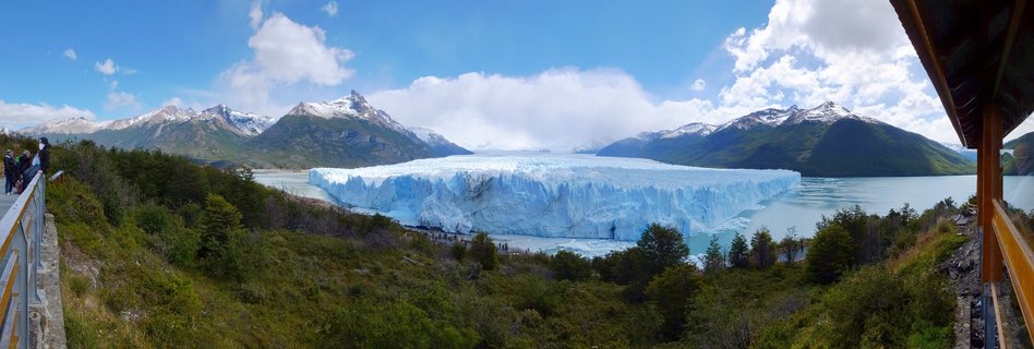 Panoramic view of Perito Moreno Glacier showing massive blue ice formation extending across lake water with snow-capped mountains in background, green forest in foreground, and tourists on viewing platforms on both sides