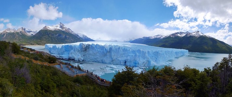 Panoramic view of Perito Moreno Glacier showing massive blue ice wall extending across turquoise lake water, with snow-capped mountains in background and wooden tourist walkways with visitors in foreground among green vegetation