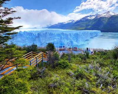 Massive blue glacier wall with wooden viewing platforms and tourists in foreground, snow-capped mountains and cloudy sky in background at Perito Moreno, Argentina